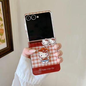 Op de afbeelding: Een doorzichtige telefoonhoes met een rood-wit ruitpatroon en een Hello Kitty-afbeelding. De hoes heeft een rode rand en de tekst "Be happy every day". Twee Hello Kitty-bedels zijn bevestigd.