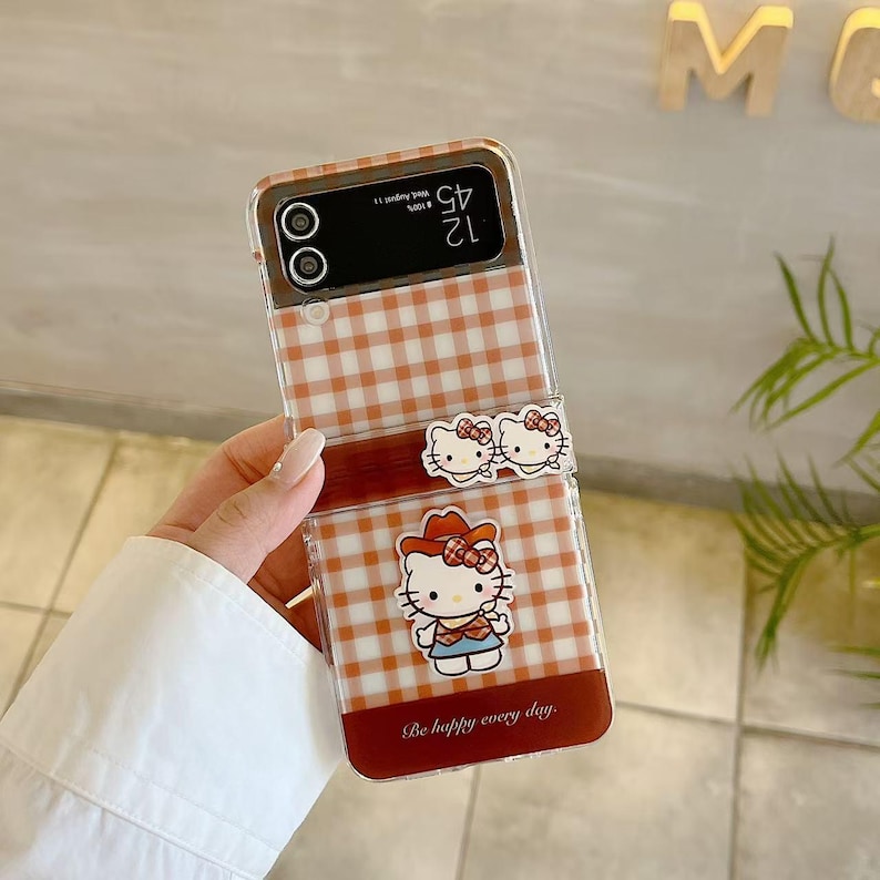 Op de afbeelding: Een doorzichtige telefoonhoes met een bruin en wit ruitpatroon. De hoes heeft een Hello Kitty-ontwerp met een rode strik en de tekst "Be happy every day."