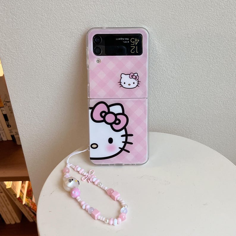 May include: Pink phone case with a Hello Kitty design. The case has a checkered pattern and a wrist strap with pink beads and a Hello Kitty charm. The phone case is open, showing the phone's screen.
