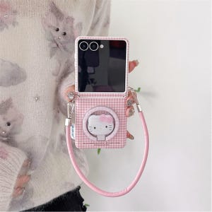 Puede incluir: Funda de teléfono a cuadros rosas y blancos con un diseño de Hello Kitty. La funda tiene una correa rosa y un diseño circular con la cara de Hello Kitty. La funda del teléfono está diseñada para abrirse y cerrarse.
