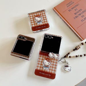 Op de afbeelding: Transparante telefoonhoesjes met een rood-wit geruit patroon en een Hello Kitty-ontwerp. De hoesjes hebben de tekst "Be happy every day". Een boek en een kralenketting met een Hello Kitty-bedel zijn ook aanwezig.