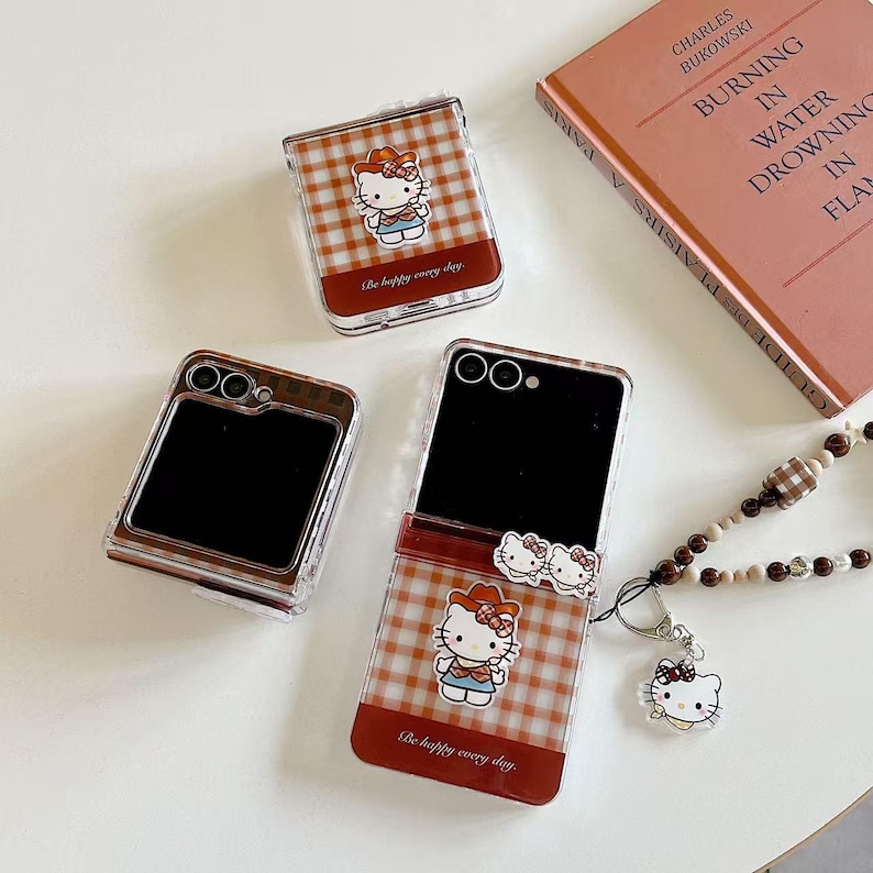 Op de afbeelding: Transparante telefoonhoesjes met een rood-wit geruit patroon en een Hello Kitty-ontwerp. De hoesjes hebben de tekst "Be happy every day". Een boek en een kralenketting met een Hello Kitty-bedel zijn ook aanwezig.