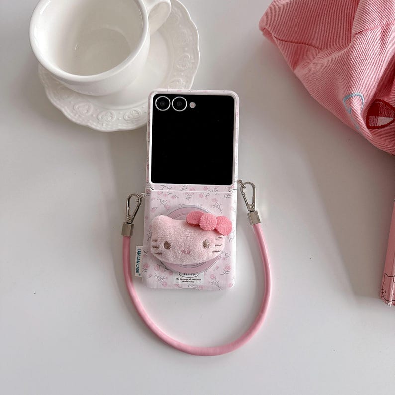 May include: Pink phone case with a Hello Kitty design, featuring a plush face and bow. The case has a matching pink strap and silver hardware. The phone screen is black. A white teacup and saucer are in the background.