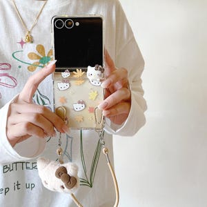 May include: Clear phone case with a Hello Kitty design, featuring cartoon characters, autumn leaves, and a plush charm. The case has a strap for carrying. The phone case is designed for a flip phone.