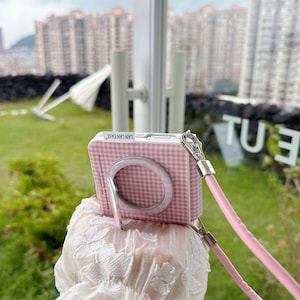 May include: A pink and white chequered phone case with a matching ring holder and a pink lanyard. The case has the text "LAN LAN CASE" printed on it. The case is designed to look like a camera.