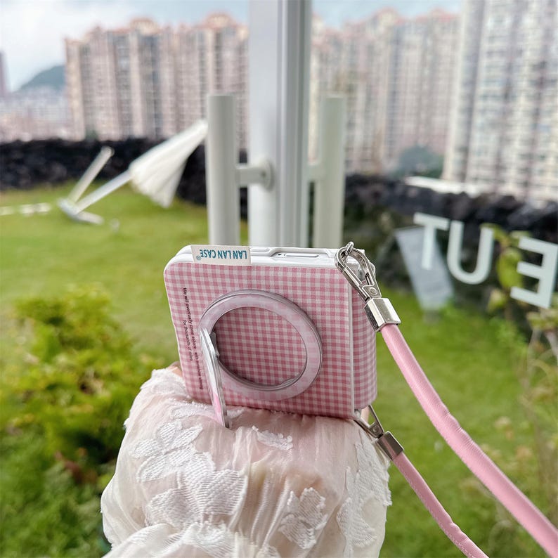 May include: A pink and white chequered phone case with a matching ring holder and a pink lanyard. The case has the text "LAN LAN CASE" printed on it. The case is designed to look like a camera.