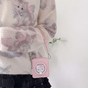 May include: A pink and white gingham pattern crossbody bag featuring a Hello Kitty design. The bag has a pink strap and silver-coloured hardware. The bag is held by a person wearing a white sweater with a floral pattern.
