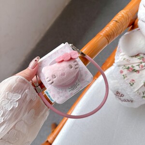 May include: A pink phone grip featuring a plush Hello Kitty design with a pink bow. The grip is attached to a pink lanyard. The packaging has a floral pattern and the text "LAN LAN CASE".