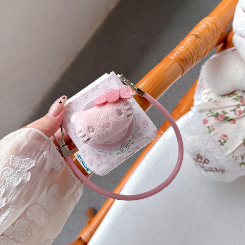 May include: A pink phone grip featuring a plush Hello Kitty design with a pink bow. The grip is attached to a pink lanyard. The packaging has a floral pattern and the text "LAN LAN CASE".