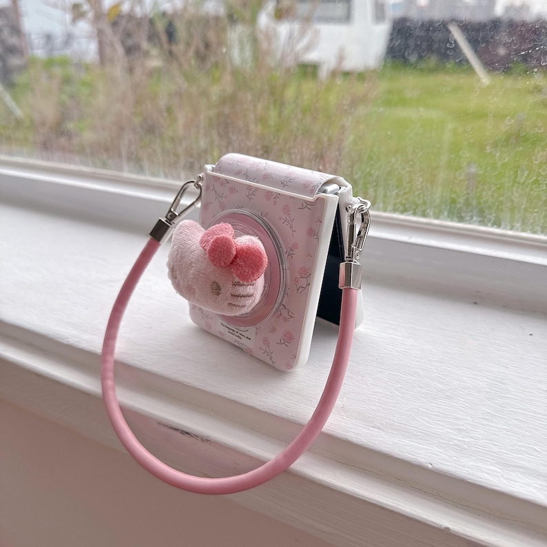 May include: A pink and white camera case featuring a Hello Kitty plushie. The case has a floral pattern and a pink strap. The plushie has a pink bow and is attached to the front of the case.