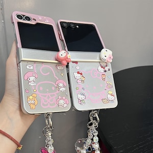 May include: Two pink and silver phone cases with cartoon character designs. The cases are open, revealing the phone screens. Each case has a charm attached to the bottom. The cases feature pink accents and cartoon character illustrations.