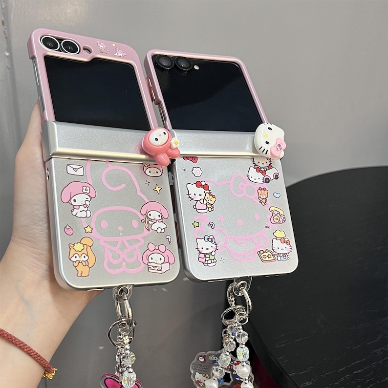 May include: Two pink and silver phone cases with cartoon character designs. The cases are open, revealing the phone screens. Each case has a charm attached to the bottom. The cases feature pink accents and cartoon character illustrations.
