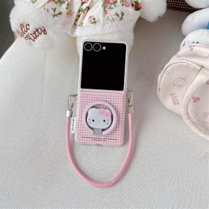 May include: A pink and white checked phone case with a Hello Kitty design. It has a matching pink strap and silver-coloured metal hardware. The phone case is open, showing the phone's screen and camera.