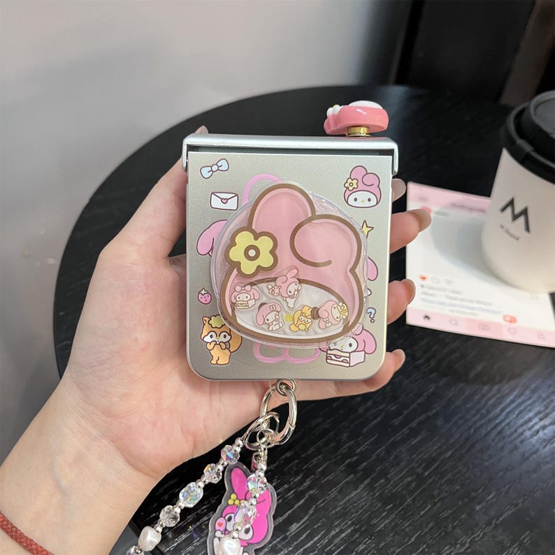May include: A silver-colored, rectangular case with a pink and yellow cartoon design. The case has a clear, rounded plastic overlay with cartoon characters inside. A pink and white button is on top, and a keychain with a pink charm is attached.