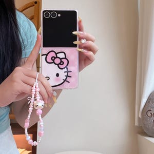 May include: A pink and black phone case with a Hello Kitty design. The case has a pink bow and face on the bottom half and a black top half. A pink and white beaded phone charm with a Hello Kitty charm is attached.