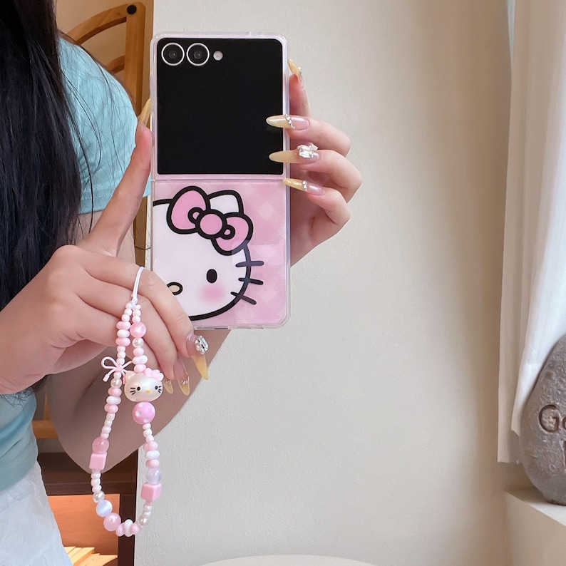 May include: A pink and black phone case with a Hello Kitty design. The case has a pink bow and face on the bottom half and a black top half. A pink and white beaded phone charm with a Hello Kitty charm is attached.