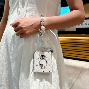 May include: White and black Hello Kitty phone case with a pearl and bow wrist strap. The case features a quilted pattern and a cartoon image of Hello Kitty wearing a hat. The wrist strap includes pearls and decorative elements.