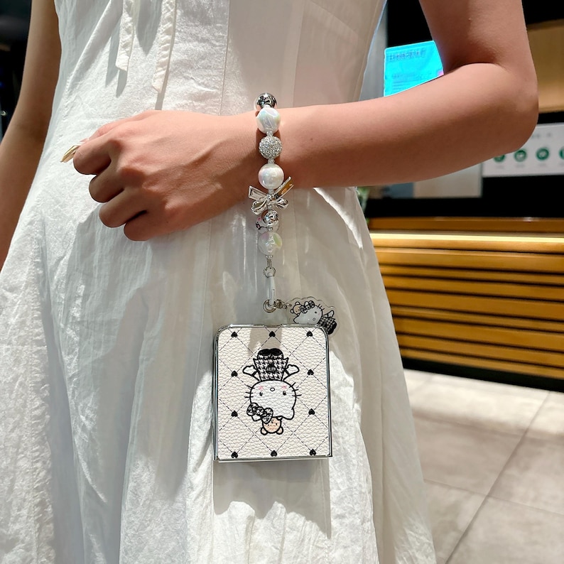 May include: White and black Hello Kitty phone case with a pearl and bow wrist strap. The case features a quilted pattern and a cartoon image of Hello Kitty wearing a hat. The wrist strap includes pearls and decorative elements.