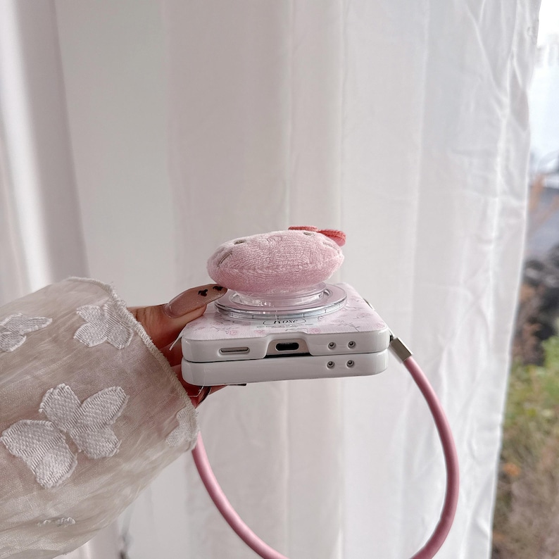 May include: A pink phone case with a floral pattern and a fluffy, circular attachment. The case has a matching pink strap. The attachment has a small red bow.