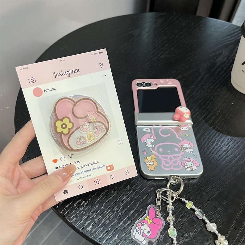 May include: Pink and silver phone case with cartoon character designs, a matching phone grip, and a keychain with a pink charm. The phone case is open, revealing the phone's screen. The phone grip is in a clear case.