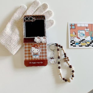 Op de afbeelding: Een doorzichtige telefoonhoes met een bruin en wit geruit patroon met een Hello Kitty-afbeelding. De hoes heeft een bijpassende sleutelhanger met een kralenband. Er is ook een kleine kaart met een Hello Kitty-illustratie.