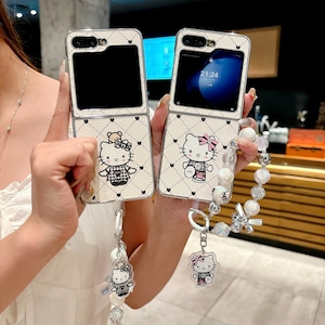 May include: Two white phone cases with a Hello Kitty design. The cases are open, revealing the phone screens. Each case has a strap with a Hello Kitty charm and pearl beads. The cases have a quilted pattern with black hearts.