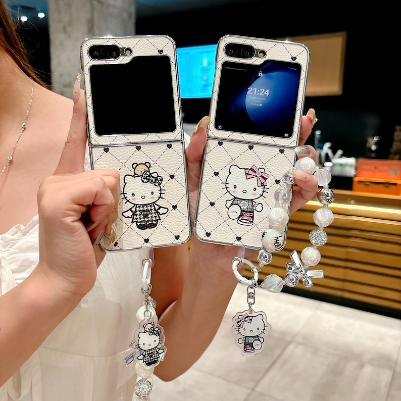 May include: Two white phone cases with a Hello Kitty design. The cases are open, revealing the phone screens. Each case has a strap with a Hello Kitty charm and pearl beads. The cases have a quilted pattern with black hearts.