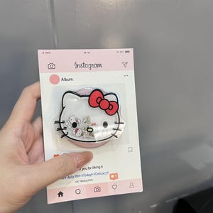 May include: A Hello Kitty phone grip with a red bow, displayed on a pink Instagram-themed card. The grip is clear with small decorative elements inside. The card has the Instagram logo and the text "Album."