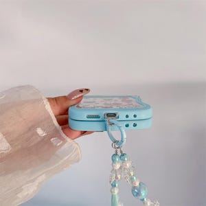 May include: A light blue phone case with a cartoon design, featuring a matching blue ring and a beaded charm. The charm includes iridescent and blue beads. The case is held by a hand with painted nails.
