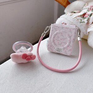May include: A pink baby pacifier with a plush bow and a matching floral-patterned case with a pink strap. The case has a floral design and the word "Rose" printed on it. The pacifier is next to the case.