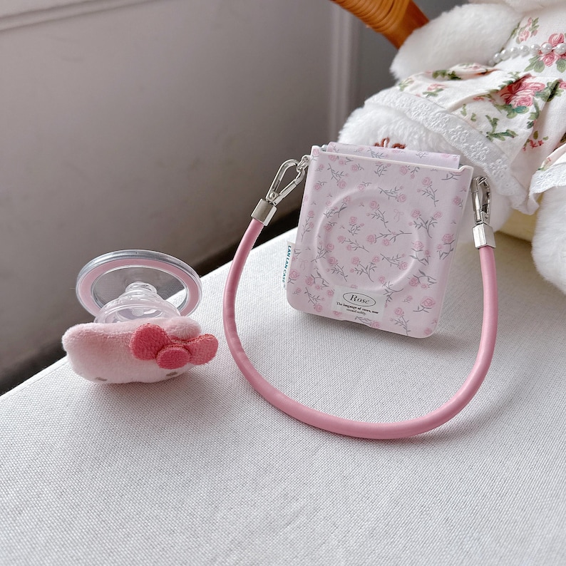 May include: A pink baby pacifier with a plush bow and a matching floral-patterned case with a pink strap. The case has a floral design and the word "Rose" printed on it. The pacifier is next to the case.