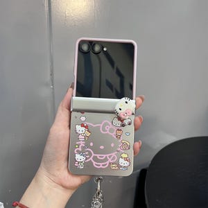 May include: A pink and silver phone case featuring a Hello Kitty design. The case has a pink border and a silver back with a pink outline of Hello Kitty and other characters. A 3D Hello Kitty charm is attached to the top.