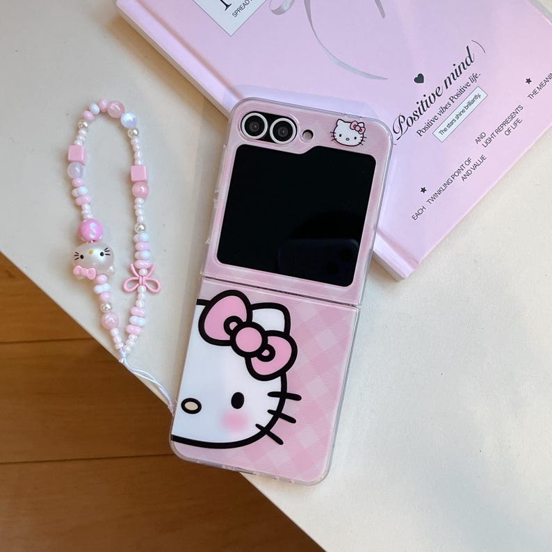 May include: A pink flip phone case with a Hello Kitty design, bow, and checkered pattern. A matching beaded phone charm with a Hello Kitty charm and pink beads is also visible. The phone case has the text "Positive mind".