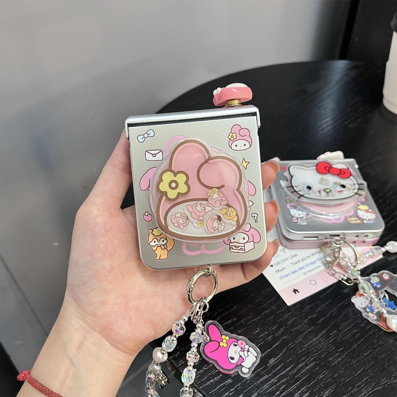 May include: A silver-colored, rectangular compact mirror with a pink, cartoon-style design. The mirror features a pink, flower-shaped button and a clear, bubble-like section with small character figures. A keychain with a pink character charm is attached.