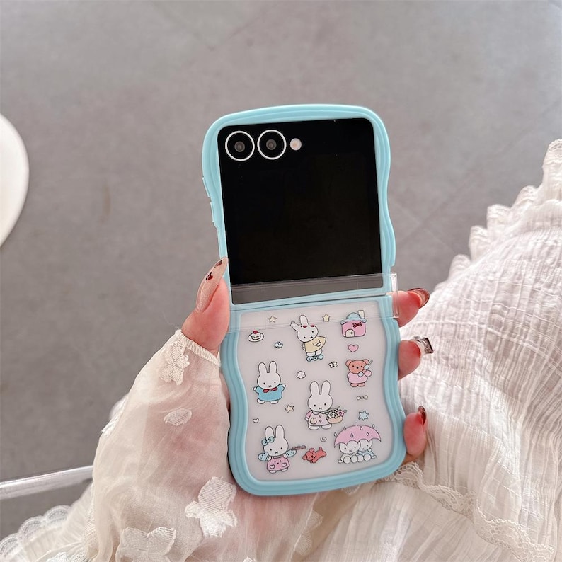 May include: A light blue phone case with a flip design. The case features a cartoon design of bunnies, bears, and other cute illustrations. The case is designed to fit a flip phone and has a protective bumper.