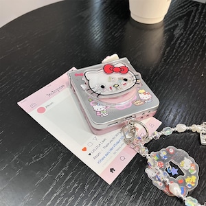 May include: A silver and pink Hello Kitty-themed camera case with a clear plastic top. The case features a red bow and other Hello Kitty designs. A pink Instagram-style card and a beaded charm are also visible.