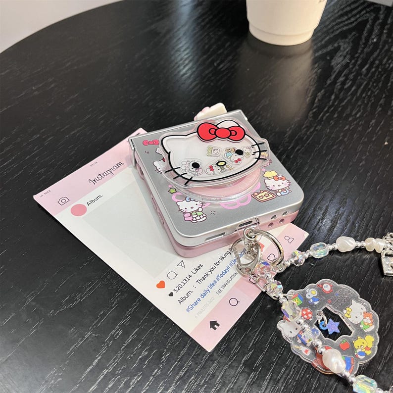 May include: A silver and pink Hello Kitty-themed camera case with a clear plastic top. The case features a red bow and other Hello Kitty designs. A pink Instagram-style card and a beaded charm are also visible.