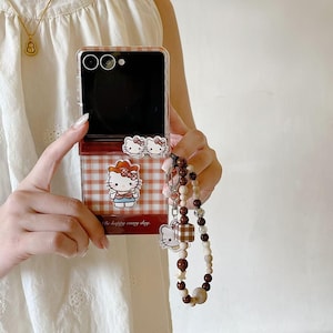 Op de afbeelding: Een doorzichtige telefoonhoes met een rood-wit geblokt patroon en een Hello Kitty-ontwerp. Het bevat een bijpassende kralenketting met een Hello Kitty-bedel en een geblokte kubus. De tekst "Be happy every day" is zichtbaar.