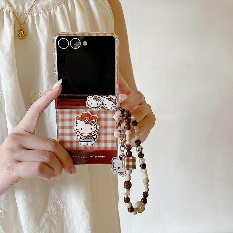 Op de afbeelding: Een doorzichtige telefoonhoes met een rood-wit geblokt patroon en een Hello Kitty-ontwerp. Het bevat een bijpassende kralenketting met een Hello Kitty-bedel en een geblokte kubus. De tekst "Be happy every day" is zichtbaar.
