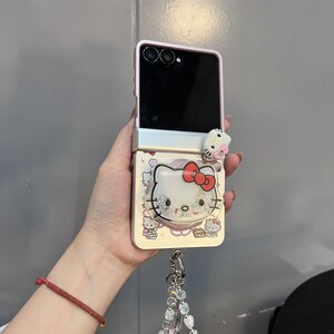 May include: A pink and white flip phone case featuring a Hello Kitty design. The case has a 3D Hello Kitty figure and a decorative charm. The phone is open, revealing the black screen.