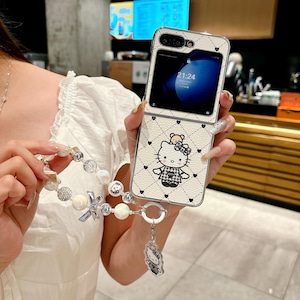 May include: A white and black patterned phone case featuring a Hello Kitty design. The case has a diamond pattern with small black hearts. A matching beaded wrist strap with a Hello Kitty charm is attached.