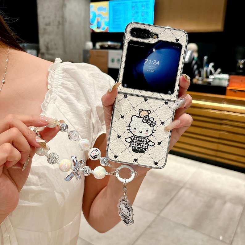 May include: A white and black patterned phone case featuring a Hello Kitty design. The case has a diamond pattern with small black hearts. A matching beaded wrist strap with a Hello Kitty charm is attached.