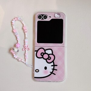 May include: Pink foldable phone case with a Hello Kitty design. The case has a pink and white checked pattern. A beaded wrist strap with pink and white beads and a Hello Kitty charm is attached.