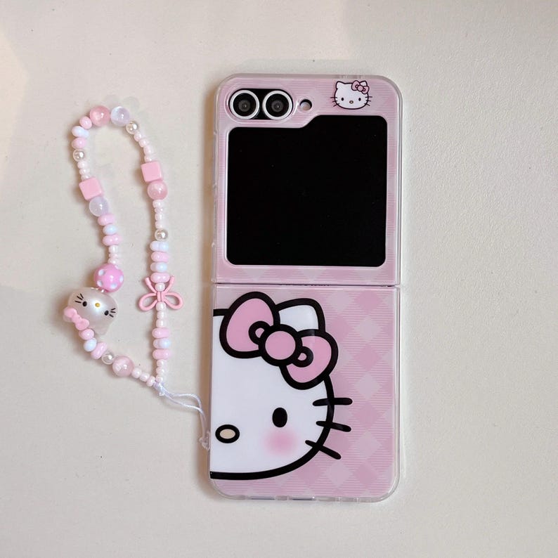 May include: Pink foldable phone case with a Hello Kitty design. The case has a pink and white checked pattern. A beaded wrist strap with pink and white beads and a Hello Kitty charm is attached.