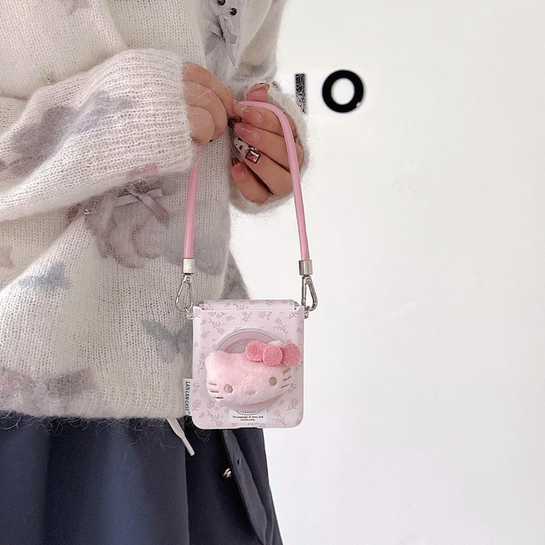 May include: A pink crossbody bag featuring a Hello Kitty design with a plush face and bow. The bag has a floral pattern and a pink strap. The bag is held by a person wearing a white sweater.