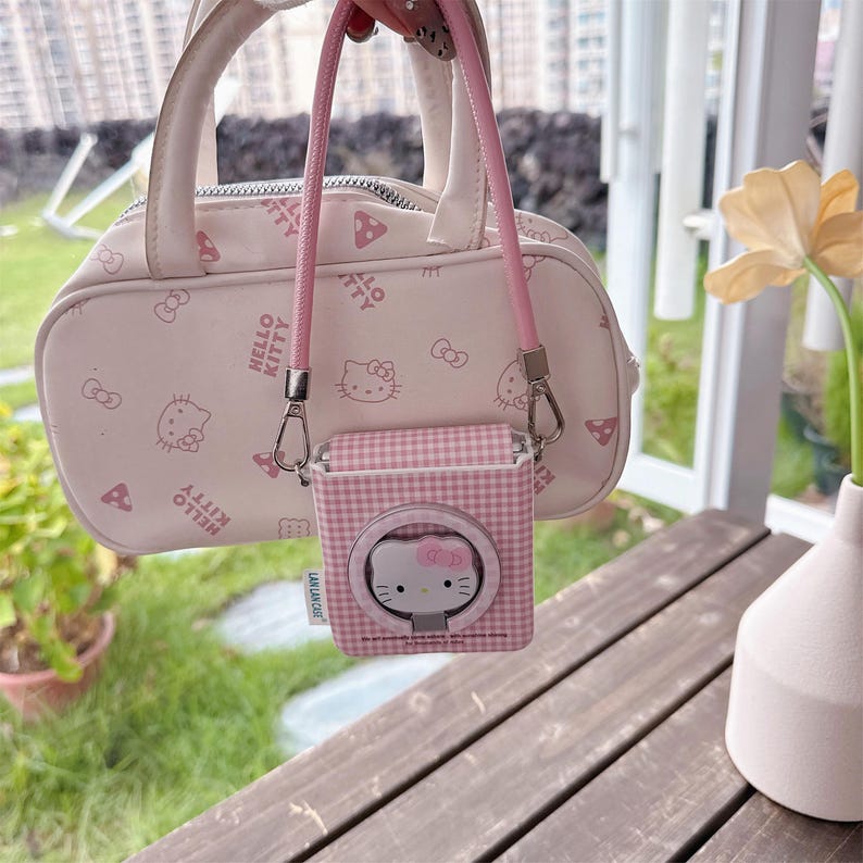 May include: A pale pink Hello Kitty handbag and AirPod case. The handbag has a top handle and a pink strap. The AirPod case is square with a Hello Kitty design and a ring holder. The background includes a wooden table and a flower.