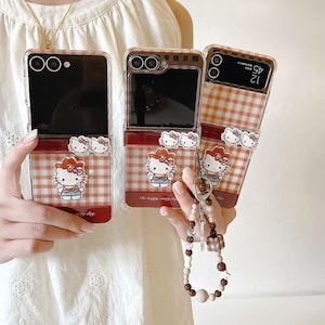 Op de afbeelding: Transparante telefoonhoesjes met een rood-wit ruitpatroon en een Hello Kitty-ontwerp. De hoesjes hebben een rode rand en een Hello Kitty-bedel. De hoesjes zijn open en tonen de schermen van de telefoons.