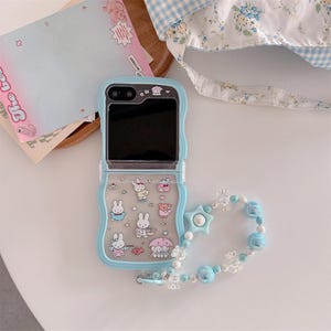 May include: A light blue phone case with a clear, patterned design featuring cartoon bunnies and other characters. The case has a matching beaded wrist strap with blue and clear beads and a star charm. A folded notebook and a floral tote bag are in the background.