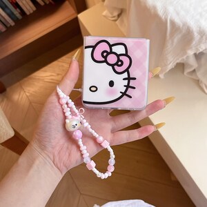 May include: A pink and white phone case featuring the Hello Kitty character, with a matching beaded charm. The case has a pink gingham pattern and a large pink bow. The charm includes a Hello Kitty face and pink beads.