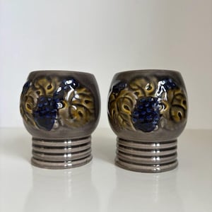 Op de afbeelding: Twee keramische bekers met een grijze basis en een afgeronde bovenkant. De bekers hebben een ontwerp van blauwe druiven en gele bladeren. De basis heeft horizontale ribbels. De bekers worden op een wit oppervlak gepresenteerd.